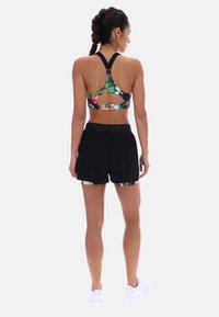 Femme aux cheveux tressés portant un brassière de sport à fleurs et un short de sport noir avec bordure fleurie, debout de dos sur un fond blanc.