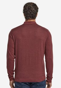 Mörkröd långärmad polo-tröja med en texturerad yta, ribbad krage och ärmslut. Stylen har en fåll som sitter vid midjan, i kombination med jeans.