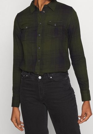 Femme portant une chemise à carreaux vert foncé et noire avec des poches à boutons-pression et un jean noir taille haute.