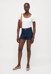 Mørkeblå denimshorts i høy midje med knappelukking. Kombinert med en tettsittende hvit T-skjorte og en brun skulderveske. Flate beige sko.