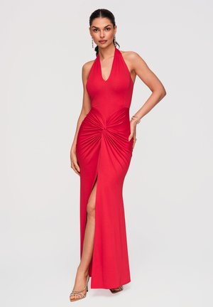 Kobieta w czerwonej wieczorowej sukni z halterem, marszczeniami z przodu i wysokim rozcięciem na udzie, ozdobiona kolczykami i bransoletkami.