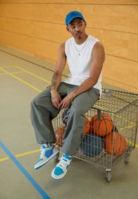 Blå kasket, ærmeløs hvid skjorte, grå cargo bukser. Fodtøj med turkise og lilla accenter. Metalvogn med basketballer i baggrunden.