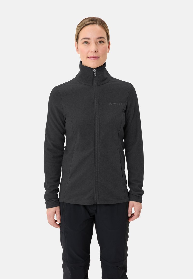 Frau trägt eine schwarze Fleecejacke mit Reißverschluss und schwarze Hose, steht vor einem einfarbigen hellen Hintergrund und blickt nach vorne.