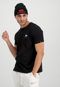 Black cotton t-shirt with short sleeves, features a small white logo on the chest, worn with a black beanie displaying red text.