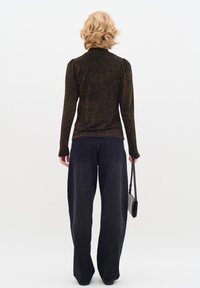Persona bionda in una maglietta a maniche lunghe con un motivo e jeans neri a gamba larga, che tiene una piccola borsa nera, in piedi su uno sfondo bianco.