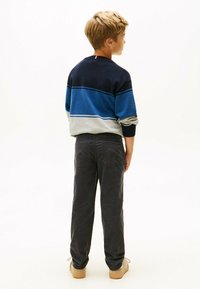 Jongen met kort blond haar staat met zijn rug naar de camera, gekleed in een marineblauwe, blauwe en grijze gestreepte trui, donkere broek en beige schoenen, tegen een witte achtergrond.