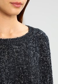 Maglione nero lavorato a maglia con una texture spessa, caratterizzato da un ampio scollo e fili di colore chiaro intrecciati per contrasto.