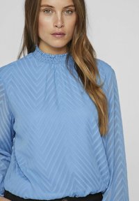 Blusa azzurra a maniche lunghe con un motivo zigzag testurizzato, scollatura arricciata e tessuto morbido e trasparente. Vita nera visibile.