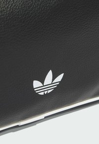 Czarna skórzana torba o teksturowanej powierzchni, z białym logo w kształcie trzylistnej koniczyny oraz trzema poziomymi paskami. Dodatki w postaci białej lamówki.