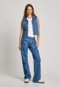Džínová vesta a wide-leg džiny v stredne modrej farbe. Vesta má bezrukávový dizajn, zapínanie na gombíky a predné vrecká. Pod ňou je biele tričko.
