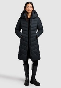 Zwarte pufferjack met quilted ontwerp, ritssluiting, grote capuchon en zijzakken. Draagt over een zwarte top en legging, gecombineerd met kniehoge laarzen.