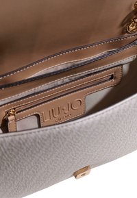 Indvendig i en lysegrå taske i struktureret læder med en lys brun foring, lynlåslomme og præget logo. Hardware i guldtoner.