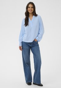 Frau mit langen dunklen Haaren, die lächelt, trägt eine hellblaue Bluse mit langen Ärmeln, blaue Jeans und schwarze Ballerinas, steht mit einer Hand in der Tasche.