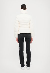 Giacca a vento in crema chiaro con colletto alto, design trapuntato e polsini a coste, abbinata a jeans neri e sneakers bianche.