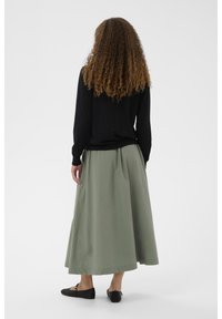 Black long-sleeve top, olive green ankle-length skirt, lightweight fabric, and black shoes with a gold accent. Curly hair visible.
