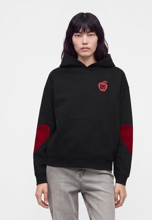 Jeune personne aux cheveux noirs et raides porte un sweat à capuche noir avec des patchs en forme de cœur rouges aux coudes et un petit logo pomme rouge sur la poitrine.