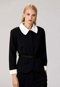 Blazer noir avec un col et des poignets blancs, doté d'une taille ceinturée et de deux poches avant. Tissu lisse avec un design structuré.