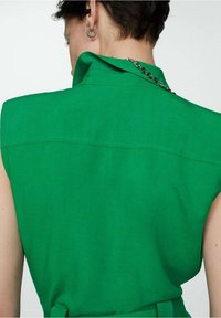 Chemise en lin vert avec un col structuré et des manches courtes, présentant une couture au dos et une ceinture à la taille, texture lisse sur toute la surface.
