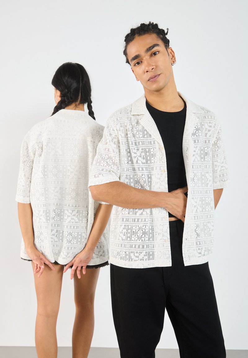 White short-sleeve shirt with intricate lace patterns, button front, relaxed fit, paired with black pants, on a male model and a female model behind.