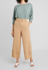 Kvinna som bär en ljusgrön texturerad långärmad topp med beige vida cropped byxor och vita öppna sandaler, stående mot enfärgad bakgrund.