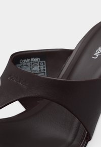 Σκούρα καφέ σανδάλι Calvin Klein slide με φαρδιά λουρίδα και ανάγλυφο λογότυπο στη λουρίδα, δείχνοντας την εσωτερική ετικέτα με το μέγεθος και τις οδηγίες φροντίδας.