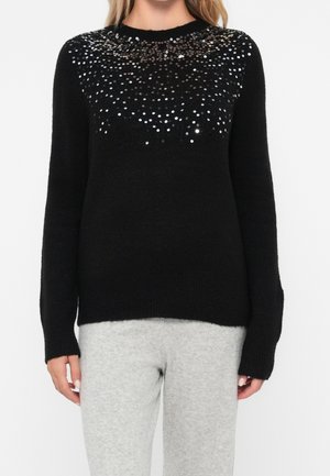 Woman wearing a black long-sleeve sweater with silver sequins on the chest and shoulders, paired with light gray pants.