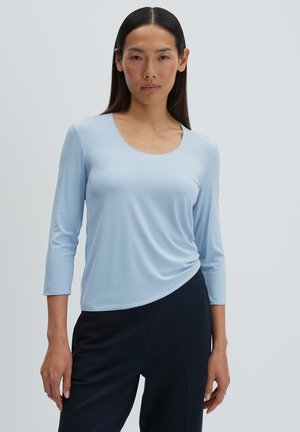 Donna con lunghi capelli scuri che indossa una maglia azzurra a maniche tre quarti e pantaloni neri su uno sfondo chiaro uniforme.