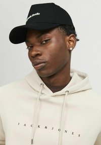 Must mustaga, millel on tekst "Studio", ja kreemivärvi kapuutsiga, millel on trükitud "JACK & JONES", varustatud paeltega ja ühtlase tekstuuriga. Suurte lähivõtte portree.