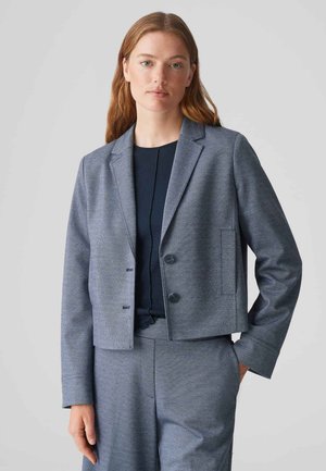 Blazer gris, court, en tissu texturé, avec revers crantés simples, deux boutons et poches latérales ; porté sur un haut sombre.