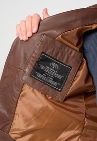 Wnętrze brązowej skórzanej kurtki z kremową satynową podszewką. Metka informuje "Skóra naturalna" oraz zawiera instrukcje pielęgnacji i szczegóły dotyczące marki.