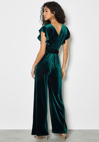 Mono de terciopelo verde profundo con mangas con volantes, diseño en V en la espalda, cintura ajustada y pantalones de pierna ancha. Textura suave con un ligero brillo.
