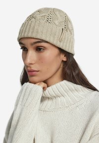 Stickad beige mössa med flätmönster och ribbad kant. Har en liten tanfärgad etikett. Bärs med en vit tjockstickad tröja som framhäver textur.