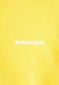 Tessuto di cotone giallo con una texture liscia, caratterizzato dal testo bianco stampato "Smilodox Sport" posizionato centralmente.