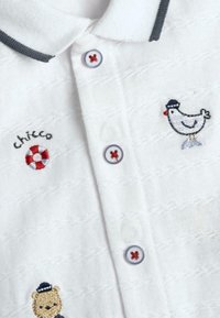 Camisa polo blanca con tejido texturizado, cuello gris, diseños bordados de un pollo y un oso, y botones con acentos rojos.