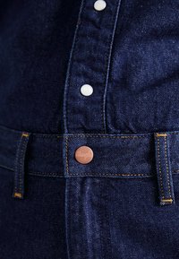 Teren din denim de culoare bleu închis, cu un prim-plan al unui buton și al plăcii cu butoane. Prezintă elemente de feronerie de ton cupru și cusături galbene în contrast.
