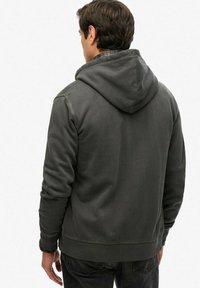 Joven vestido con una sudadera con capucha de color gris oscuro, visto desde atrás, sobre un fondo blanco liso.