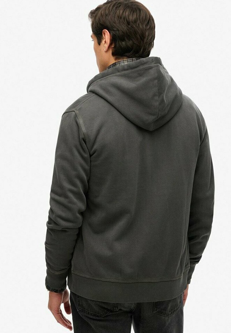 Młody mężczyzna w ciemnoszarej bluzie z kapturem, widziany od tyłu na tle jednolitego białego tła.