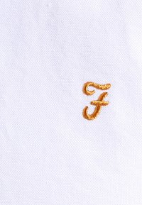 Tejido blanco con una superficie texturada que presenta una letra 'F' bordada en naranja en un estilo de escritura estilizado.