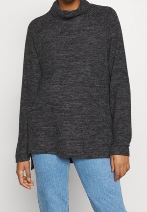 Frau trägt einen dunkelgrauen, langärmligen Pullover mit lockerem Rollkragen und hellblauer Jeans, die Hände entspannt an den Seiten.