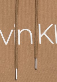 Sudadera marrón con textura acanalada, que presenta el logotipo blanco de "Calvin Klein". Tiene cordones con puntas de metal plateado para el ajuste.