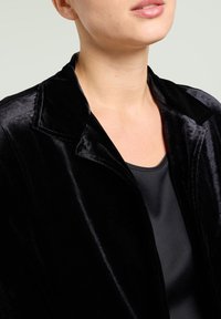 Black velvet jacket with a shawl collar, featuring a smooth texture and a relaxed fit, paired with a black satin top underneath.