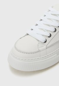 Adidași din piele albă cu talpă din cauciuc texturat, vârf rotund și șireturi plate albe. Prezintă detalii cusute și unghiuri metalice pentru șireturi.
