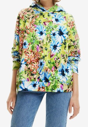Person iført en hættetrøje med blomsterprint i grønne, blå, pink og hvide toner kombineret med blå denimjeans, stående med hænder afslappet langs siden.