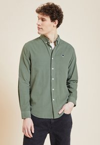 Groene button-up shirt van katoen met een gladde textuur, voorzien van een klein marineblauw boomembleem op de borst en lange mouwen.