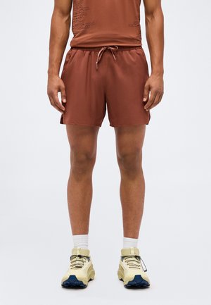 Mann trägt rostfarbene Sportshorts, weiße Socken und beige Laufschuhe mit blauen Sohlen, steht vor einem einfarbigen Hintergrund.