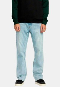 Hombre vistiendo jeans rectos de color azul claro, zapatillas negras con suelas blancas y una camiseta de manga larga negra y verde, de pie de manera relajada.