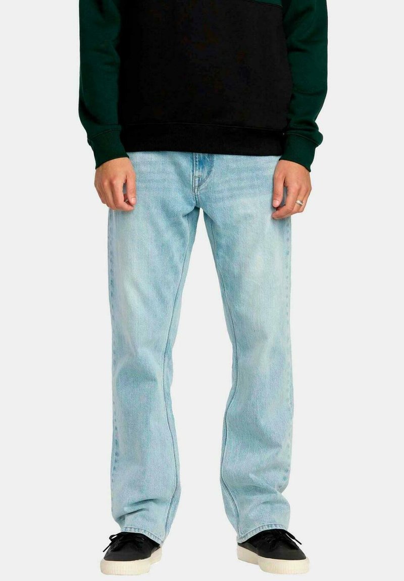 Hombre vistiendo jeans rectos de color azul claro, zapatillas negras con suelas blancas y una camiseta de manga larga negra y verde, de pie de manera relajada.