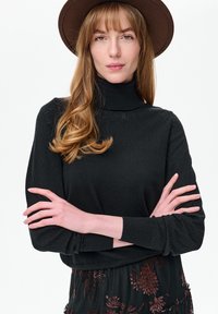 Vrouw met lang golvend haar, een bruine hoed, een zwarte coltrui en een bloemenrok, met gekruiste armen tegen een witte achtergrond.