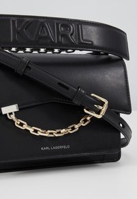 Borsa in pelle nera di Karl Lagerfeld con logo a rilievo sulla tracolla, dettaglio della catena dorata, tracolla regolabile con fibbia e chiusura a patta.