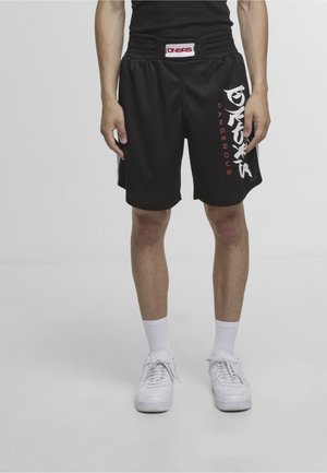 Osoba w czarnych szortach z napisem "DANGERS", białych skarpetkach i białych trampkach na jasnym, jednolitym tle.